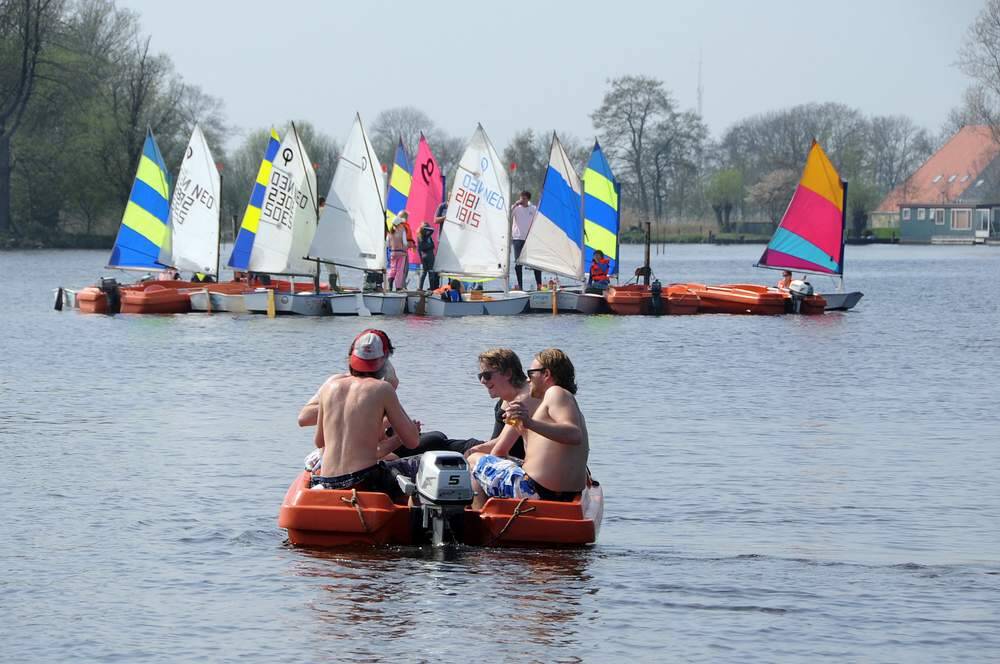 Vier jongemannen in een bootje op de Wijde Ee bij Grou. Op de achtergrond een vloot Optimistjes van zeilschool Pean in Akkrum. De leerlingen hebben pauze op een drijvend plateau, dat tijdens de zeillessen als uitvalsbasis wordt gebruikt.